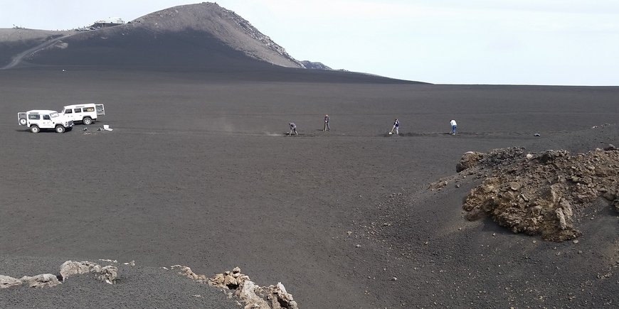 Two white off-road vehicles are parked on a grey scoria field on the volcano Etna and four people are digging the trench for the fibre optic cable with shovels.