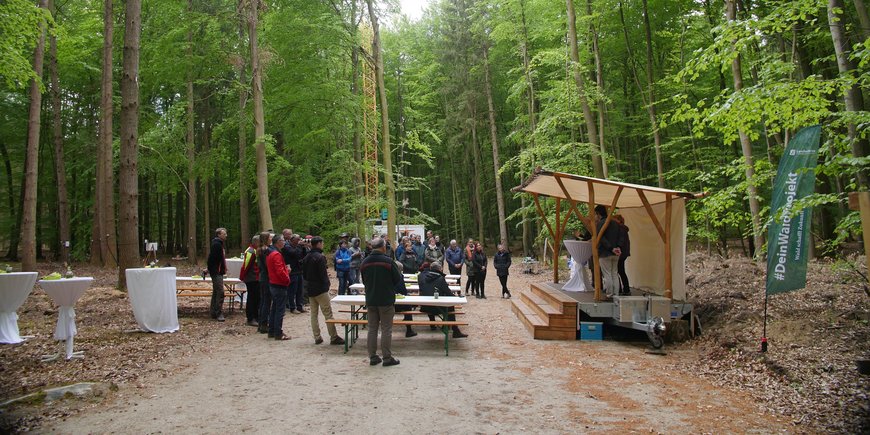 Im Wald bei Demmin wurde eine kleine Holzbühne und -tische aufgebaut. Es sind Projektverantwortliche und Politiker:innen zu sehen