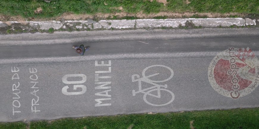 [Translate to English:] senkrechter Blick von oben auf eine Straße, auf der Straße steht geschrieben: "Tour de France Go Mantle", darunter ist ein Fahrrad abgebildet und darunter ein Logo (rot-weiß) von "Geology of Geosports"