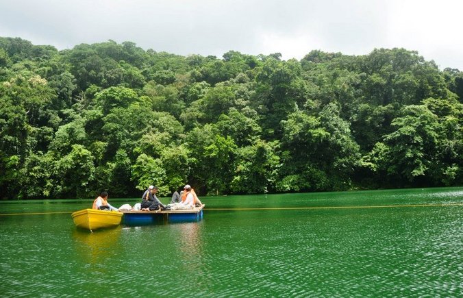 Green lake with two boats in which people are sitting. In the background green trees.