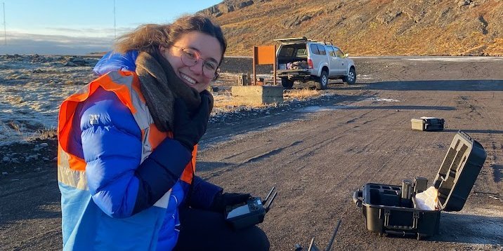 Maria Hurley on a dirt road in Iceland (dark ground), she holds a remote control for the drone in her hand, in the background an off-road vehicle, behind it a hill