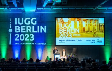 Two persons standing on stage and speaking, with the IUGG logo superimposed over her on a large screen and projected on the wall. The room is dark and many people are seated as spectators.